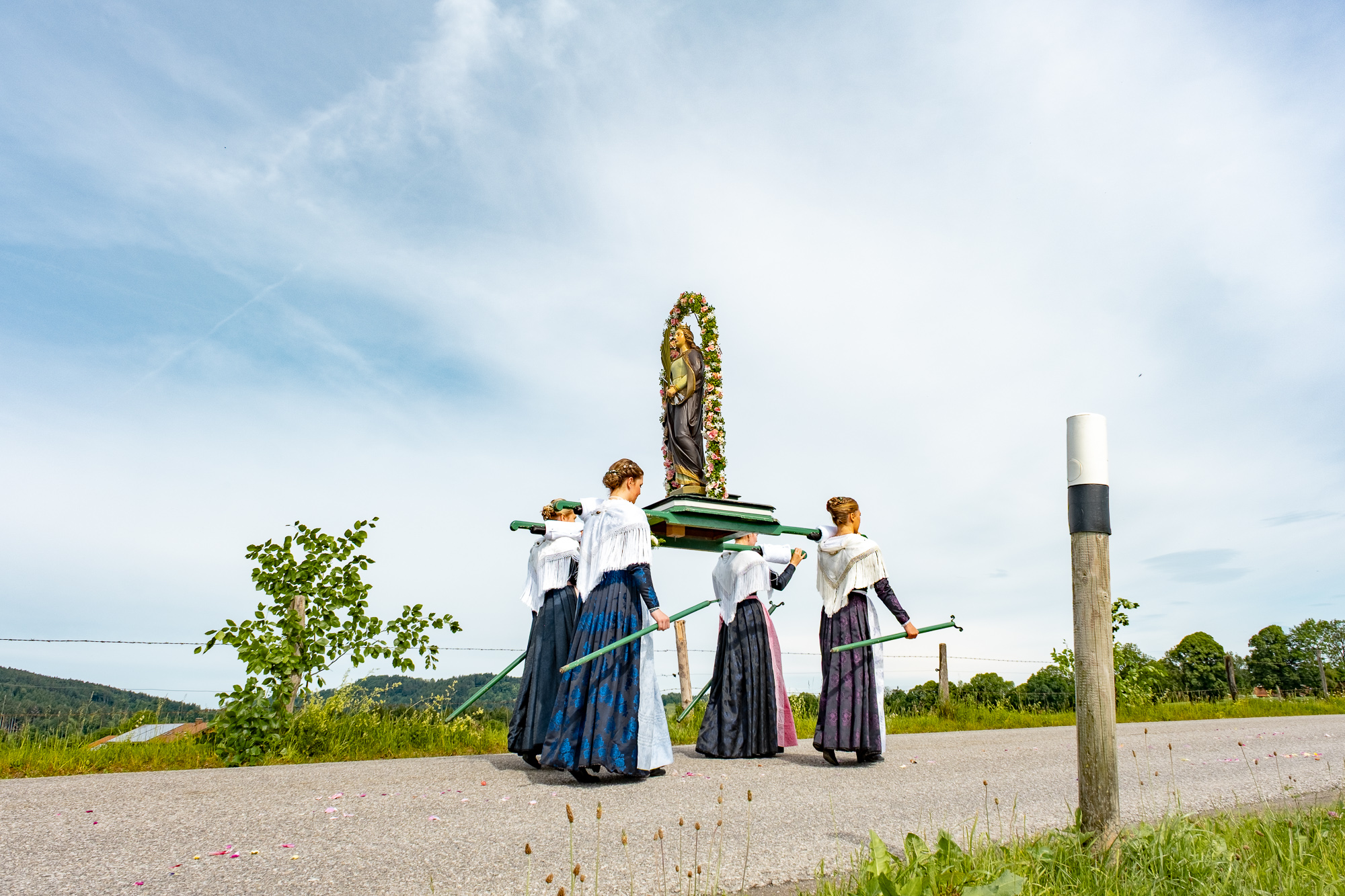 Fronleichnamsprozession in Wackersberg Fronleichnamsprozession in Wackersberg, Bayern. Vier Personen tragen eine Statue auf Stangen durch ein Feld entlang einer Strasse. Im Hintergrund sind Hügel und Bäume zu sehen. Der Himmel ist blau mit vereinzelten Wolken.